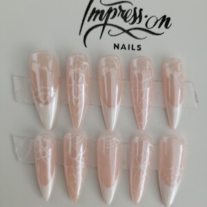 Op de afbeelding: Set van tien lange, stiletto-vormige kunstnagels met een witte tip en een delicaat wit spinnenwebontwerp op een lichtroze basis. De nagels zijn in twee rijen gerangschikt op een witte kaart met de tekst "Impress-on Nails" in zwart gedrukt.