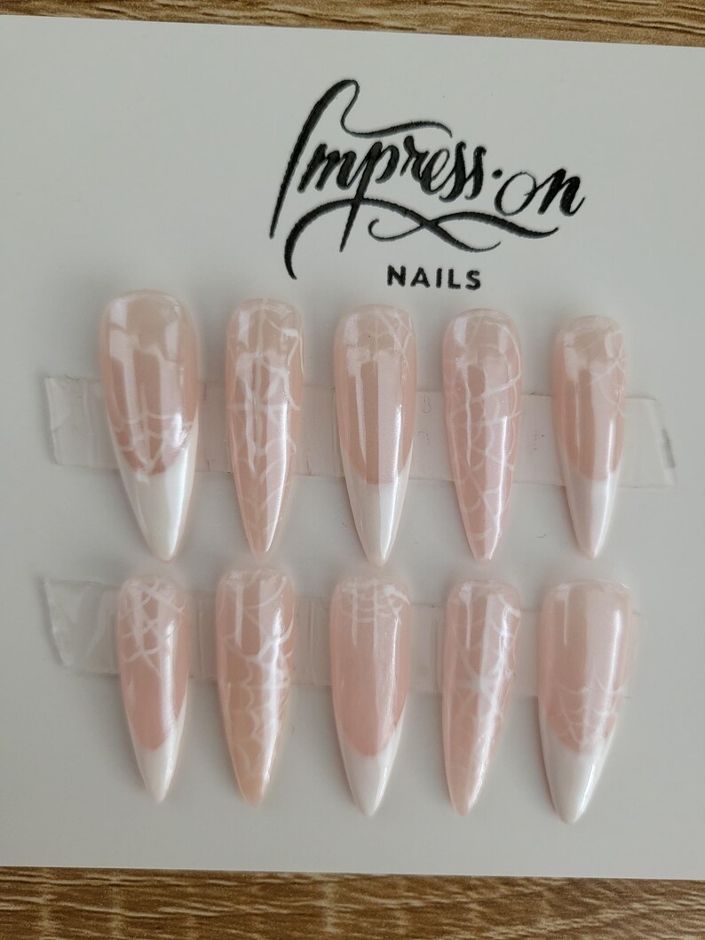 Op de afbeelding: Set van tien lange, stiletto-vormige kunstnagels met een witte tip en een delicaat wit spinnenwebontwerp op een lichtroze basis. De nagels zijn in twee rijen gerangschikt op een witte kaart met de tekst "Impress-on Nails" in zwart gedrukt.