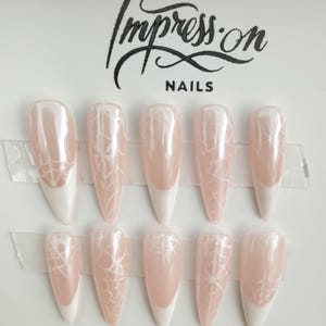Op de afbeelding: Set van tien lange, stiletto-vormige kunstnagels met een witte tip en een spinnenwebontwerp op een lichtroze basis. De nagels zijn gerangschikt in twee rijen van vijf op een witte achtergrond. De tekst "Impress.on NAILS" is bovenaan de afbeelding gedrukt.