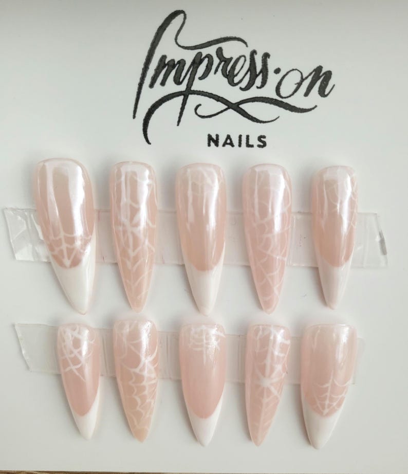 Op de afbeelding: Set van tien lange, stiletto-vormige kunstnagels met een witte tip en een spinnenwebontwerp op een lichtroze basis. De nagels zijn gerangschikt in twee rijen van vijf op een witte achtergrond. De tekst "Impress.on NAILS" is bovenaan de afbeelding gedrukt.