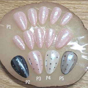 May include: A selection of false nails in shades of pink, black, white, and grey. Each nail features small, evenly spaced white or black dots. The nails are displayed on a light-coloured, oval-shaped surface.