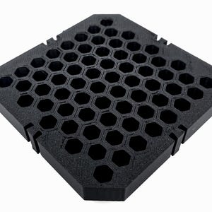 May include: A black, geometric honeycomb-patterned object. The object is square with cut-out sections on the sides. The surface is covered with a series of hexagonal holes, creating a grid-like appearance. The object is made of a matte black material.