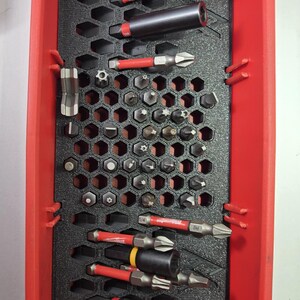 May include: A red plastic tool organizer with a black honeycomb insert. The organizer contains a variety of drill bits and screwdriver bits, including a 1/4 inch bit and a 5/16 inch bit.