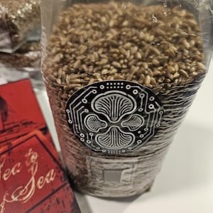 May include: A clear plastic bag filled with brown grains, possibly for growing mushrooms. A black and white sticker with a mushroom design is on the bag. A red box with the words "Tea to Sea" is visible.