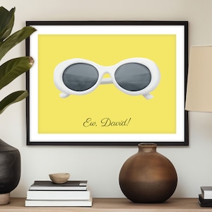 May include: A framed print with a yellow background featuring a pair of white oval sunglasses with dark lenses. The text "Ew, David!" is printed below the sunglasses.