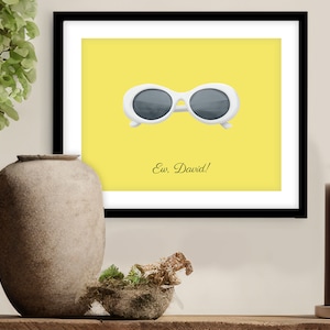 May include: A framed print with a yellow background featuring a pair of white sunglasses with a blue and gray reflective lens. The text "Ew, David!" is printed in black below the sunglasses.