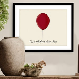 May include: Framed art print featuring a red balloon with a thin white string against a cream background. The text "We all float down here" is written in a cursive font below the balloon. The print is in a black frame.