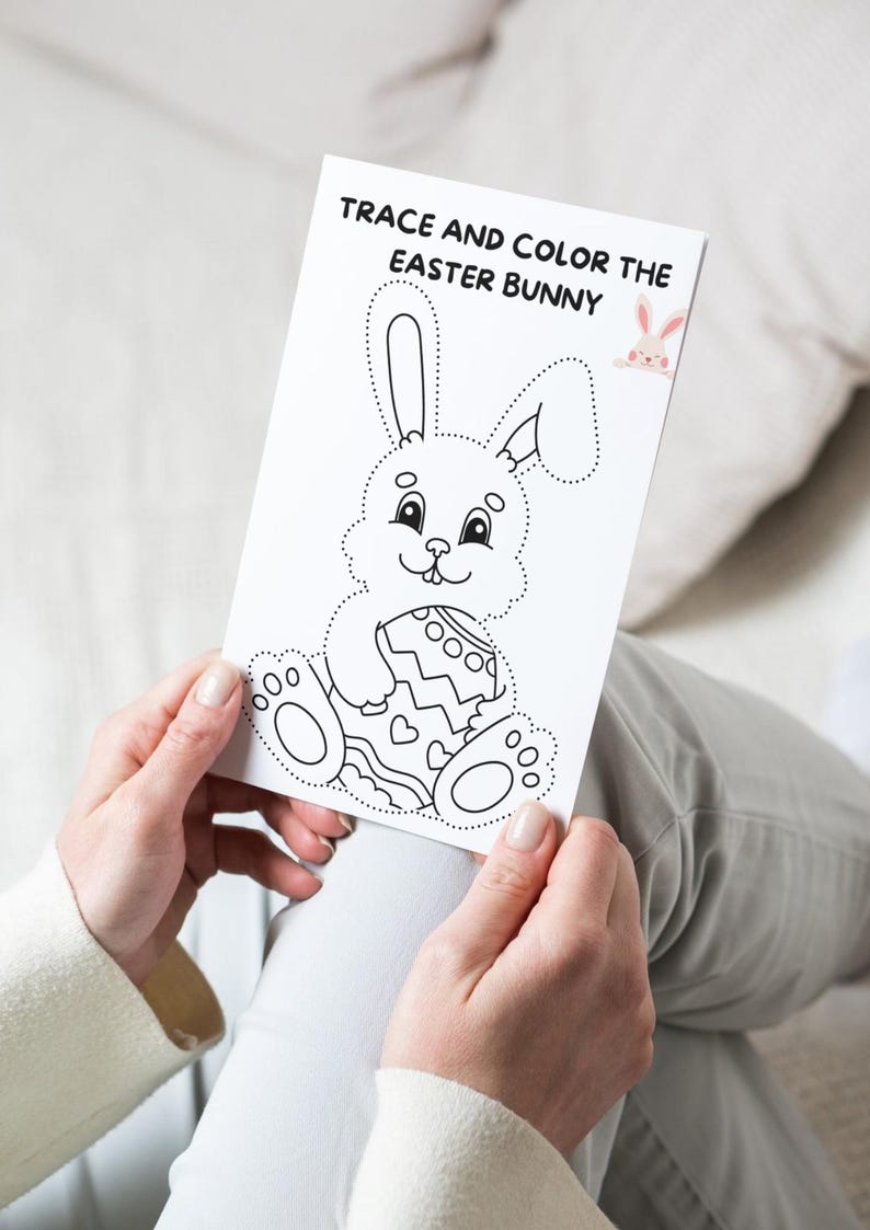 May include: A white coloring page with the text "TRACE AND COLOR THE EASTER BUNNY" features a dotted outline of a bunny holding an Easter egg. A small pink bunny graphic is in the upper right corner. The page is held by a person.