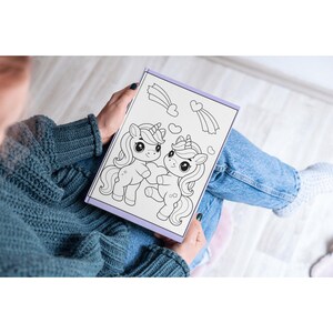 May include: A coloring book with a black and white illustration of two unicorns with hearts and shooting stars. The book is open and being held by a person.