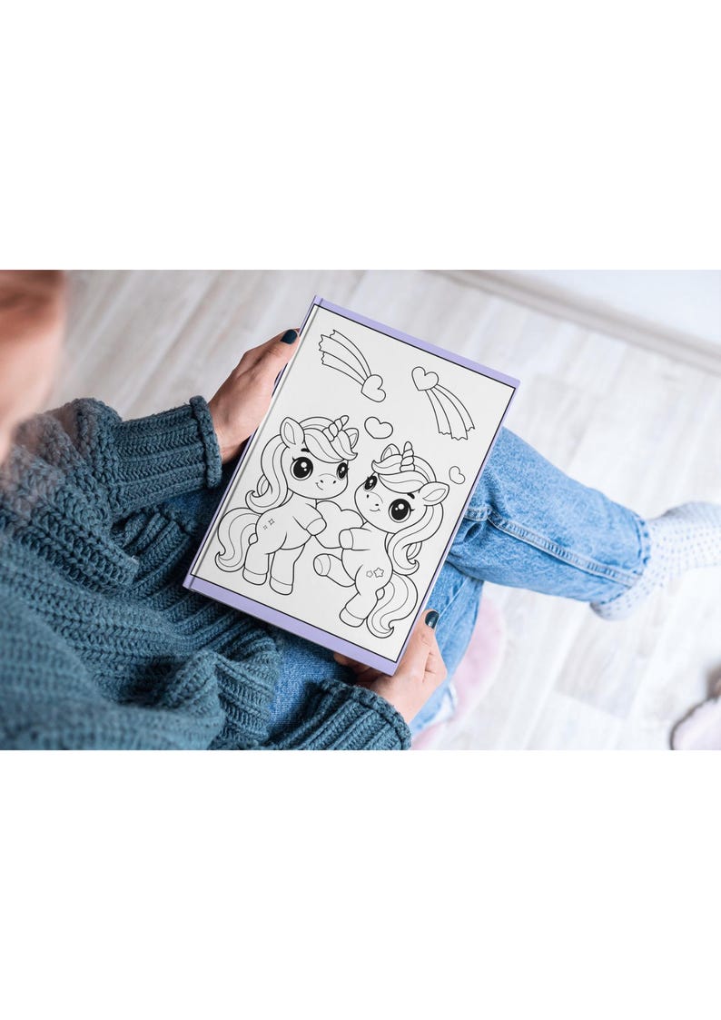 May include: A coloring book with a black and white illustration of two unicorns with hearts and shooting stars. The book is open and being held by a person.