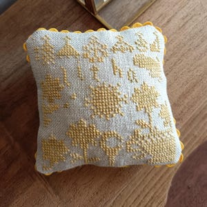 Può includere: Un piccolo cuscino quadrato bianco con un disegno ricamato giallo di alberi e fiori. Il cuscino ha un bordo giallo a festoni.