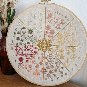 Peut inclure: Un cercle de broderie au point de croix avec un motif floral aux couleurs variées. Le motif est divisé en sections, chacune avec un motif floral et une palette de couleurs différents, dont le rose, le jaune et le vert. Le texte "betaine" est visible.