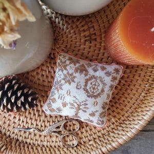 以下が含まれることがあります： 茶色の花柄刺繍が施された、クリーム色の正方形の小さなクッション。クッションは、松ぼっくりと金色のハサミが置かれた籐製のトレイの上に置かれています。