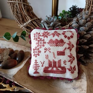 May include: A small, square, red and white embroidered pillow with a scalloped edge. The pillow features a variety of designs, including a heart, a tree, and a bird. The pillow is sitting on a wooden surface.