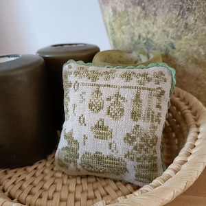 Puede incluir: Una pequeña almohada bordada en verde y blanco con un diseño floral se encuentra en una cesta tejida. La almohada tiene un borde festoneado.