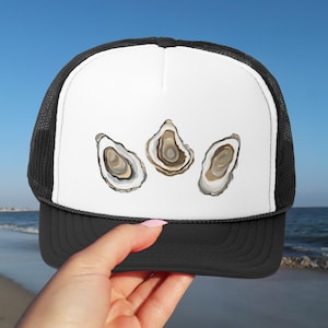 Puede incluir: Una gorra de camionero negra y blanca con un panel frontal blanco que presenta tres ilustraciones de ostras marrones y blancas.