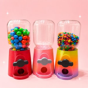 May include: Three gumball machines with clear glass globes and plastic bases. The left base is solid red, the center is a pink-to-orange gradient, and the right is a yellow-to-purple gradient. The left and right machines are filled with colorful candies.