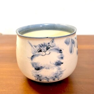 May include: A small, round ceramic cup with a cream-coloured base and a blue-painted rim. The exterior features a hand-painted illustration of a fluffy, wide-eyed cat in shades of blue, along with delicate blue leaf patterns on the side.