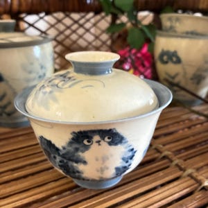 Peut inclure: Un bol à thé en céramique avec son couvercle assorti, orné d'une illustration peinte à la main en bleu et blanc représentant un chat touffu. Le bol présente une base crème claire avec un bord bleu et un petit bouton circulaire sur le couvercle.