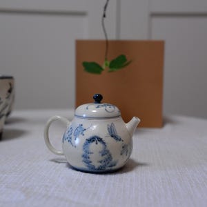 May include: A small, white ceramic teapot with blue floral and animal designs. The teapot has a curved handle, a spout, and a lid with a dark blue knob. The teapot is on a white tablecloth.