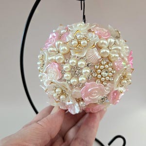 Puede incluir: Adorno esférico decorativo, adornado con adornos florales blancos y rosas, perlas de imitación y brillantes pedrería. El adorno está suspendido de un gancho de metal negro, creando una exhibición festiva y elegante.