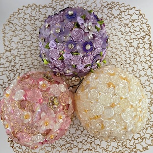 Puede incluir: Tres esferas florales decorativas en tonos morado, rosa y marfil, adornadas con diversas formas de flores, cuentas y pedrería. Las esferas descansan sobre una base circular calada de color dorado. Las esferas son probablemente para bodas o decoración del hogar.