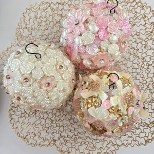 Op de afbeelding: Drie decoratieve kerstballen. Elke bol is bedekt met witte, roze en gouden bloemversieringen, parels en strass. De ballen hebben zwarte metalen haken om op te hangen en worden tentoongesteld op een gouden draadgaas.