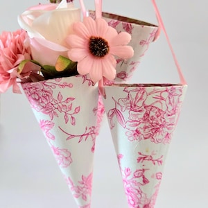 Pode incluir: Três cones florais suspensos com padrões florais rosa e fitas rosa. Os cones são preenchidos com flores rosa e brancas. Os cones são suspensos por uma mão, criando uma exibição decorativa.