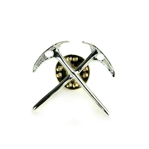 May include: Silver ice axe pin with a crossed design. The pin has a circular back with a gold finish.