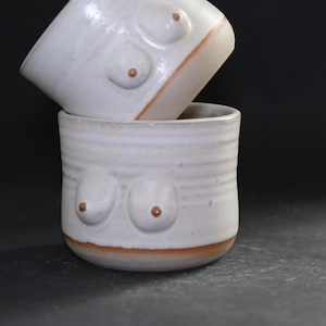 Peut inclure: Deux tasses en céramique blanche faites à la main empilées l'une sur l'autre. Chaque tasse présente une paire de formes arrondies en relief en forme de seins avec des détails de tétons bruns. La base de chaque tasse a une bande marron.