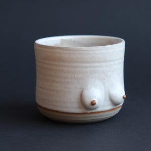 Pode incluir: Uma caneca cilíndrica de cerâmica com esmalte esbranquiçado salpicado e uma superfície texturizada com nervuras. O exterior apresenta duas protuberâncias arredondadas em relevo com pequenos pontos castanhos distintos no centro e uma fina faixa castanha na base.