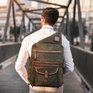 Può includere: Una persona indossa una camicia bianca e uno zaino in tela verde con dettagli in pelle marrone. Lo zaino ha una grande tasca frontale e diversi scomparti con cerniera.