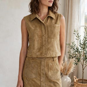 May include: A tan suede two-piece set consisting of a sleeveless button-down collared vest with a single chest pocket and a matching mini skirt featuring a front button closure and two patch pockets, both crafted from a soft, textured material.