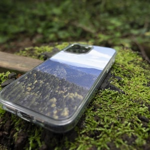 May include: A clear phone case with a mountain landscape printed on the back. The case is lying on a mossy surface.