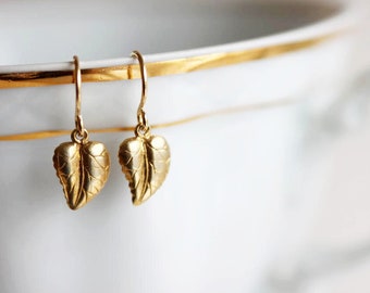 May include: A pair of gold leaf earrings with a delicate design. The earrings are hanging from a white surface.