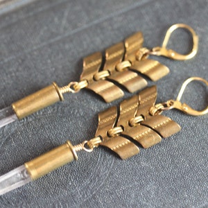 May include: A pair of gold earrings with a clear quartz crystal and a brass arrow design. The earrings are made with a bullet casing and a hook closure.