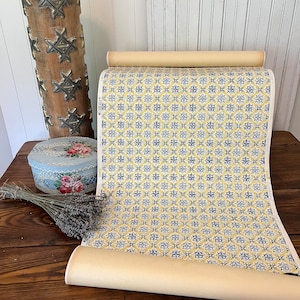 May include: A roll of yellow and blue patterned wallpaper with a repeating geometric design. The wallpaper is rolled up and sitting on a wooden table.