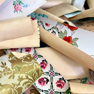 May include: A collection of vintage wallpaper scraps in various patterns and colors. The scraps include floral designs in pink, red, and green, along with gold and cream-colored patterns. Some pieces are rolled, while others are flat.