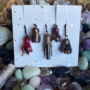 Puede incluir: Cinco pequeños colgantes de cristal en tonos rojos, marrones y dorados cuelgan de una tarjeta blanca con detalles de estrellas doradas. Los colgantes se exhiben sobre un fondo de varias piedras preciosas de colores y una cesta tejida.