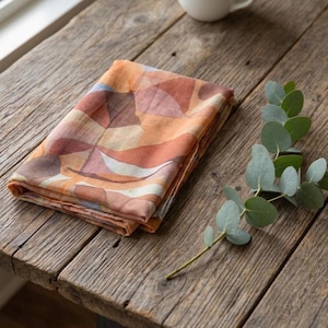 May include: A folded rectangular textile featuring an abstract pattern of overlapping shapes in shades of terracotta, burnt orange, cream, and muted blue. The fabric has a soft, lightweight appearance with visible weave texture and folded edges.