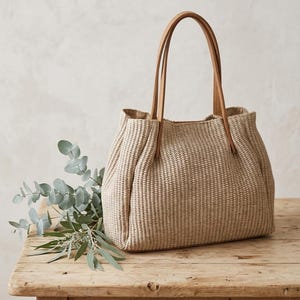 Könnte beinhalten: Eine strukturierte Tragetasche aus gewebtem beigefarbenem Material mit texturierter Oberfläche. Die Tasche verfügt über zwei schmale, hellbraune Griffe an der Oberseite. Das Design umfasst einen rechteckigen Boden und vertikale Falten an den Seiten.