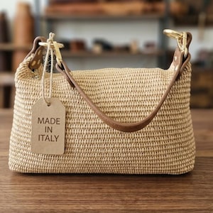 May include: A beige woven handbag with a rectangular shape, featuring a brown leather handle attached with gold-tone metal hardware. A rectangular cardboard tag hangs from the handle, with the text MADE IN ITALY printed in black ink.