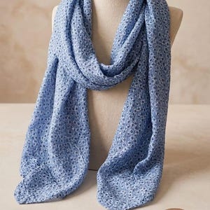 May include: A light blue scarf with a delicate floral pattern is draped over a mannequin. The scarf is made of a lightweight fabric. A wooden spool and a round sticker with text and a sunflower are in the background.