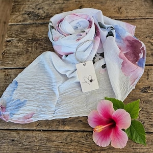 May include: A white scarf with a watercolor floral pattern in shades of pink, purple, and blue. The scarf has a crinkled texture and features a rectangular white tag attached with string, displaying a heart icon and the text MADE IN ITALY.