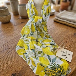 May include: A lightweight scarf with a lemon print in yellow and white, draped over a mannequin head. The scarf has a tag that says "made in Italy". A sprig of yellow flowers is next to the tag.