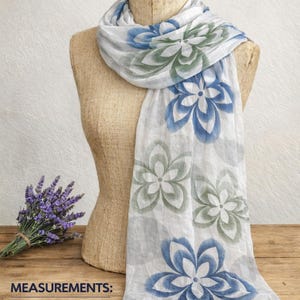 May include: A white scarf featuring a repeating floral pattern in shades of blue and green. The scarf measures 145 cm in length and 80 cm in width. The fabric has a lightweight appearance and is draped over a mannequin torso.