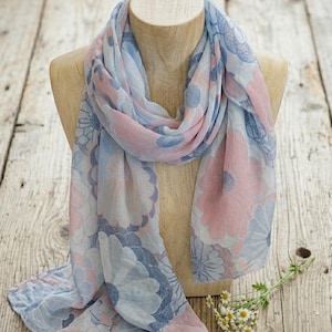 May include: A lightweight scarf featuring a large-scale floral pattern in soft shades of pastel pink, light blue, and muted periwinkle. The fabric is draped around a wooden mannequin neck, displaying a repeating circular flower motif throughout.