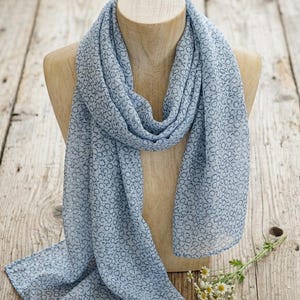 May include: A light blue scarf featuring a repeating pattern of small, dark gray circular outlines. The scarf is draped over a wooden mannequin bust, showcasing its lightweight, semi-sheer fabric and consistent print across the entire surface.