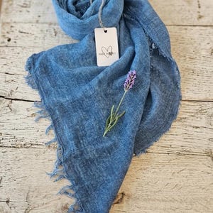 May include: A blue scarf with a frayed edge and a soft, textured appearance. A white rectangular tag is attached to the fabric, featuring a hand-drawn heart icon and the text "MADE IN ITALY". A single sprig of purple lavender rests on the scarf.