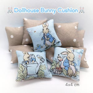 May include: Dollhouse bunny cushions, each measuring 4x4 cm. The cushions feature illustrations of a bunny in a blue jacket. Some cushions are light blue with bunny illustrations, while others are tan with white polka dots. The text "Dollhouse Bunny Cushion" is at the top.